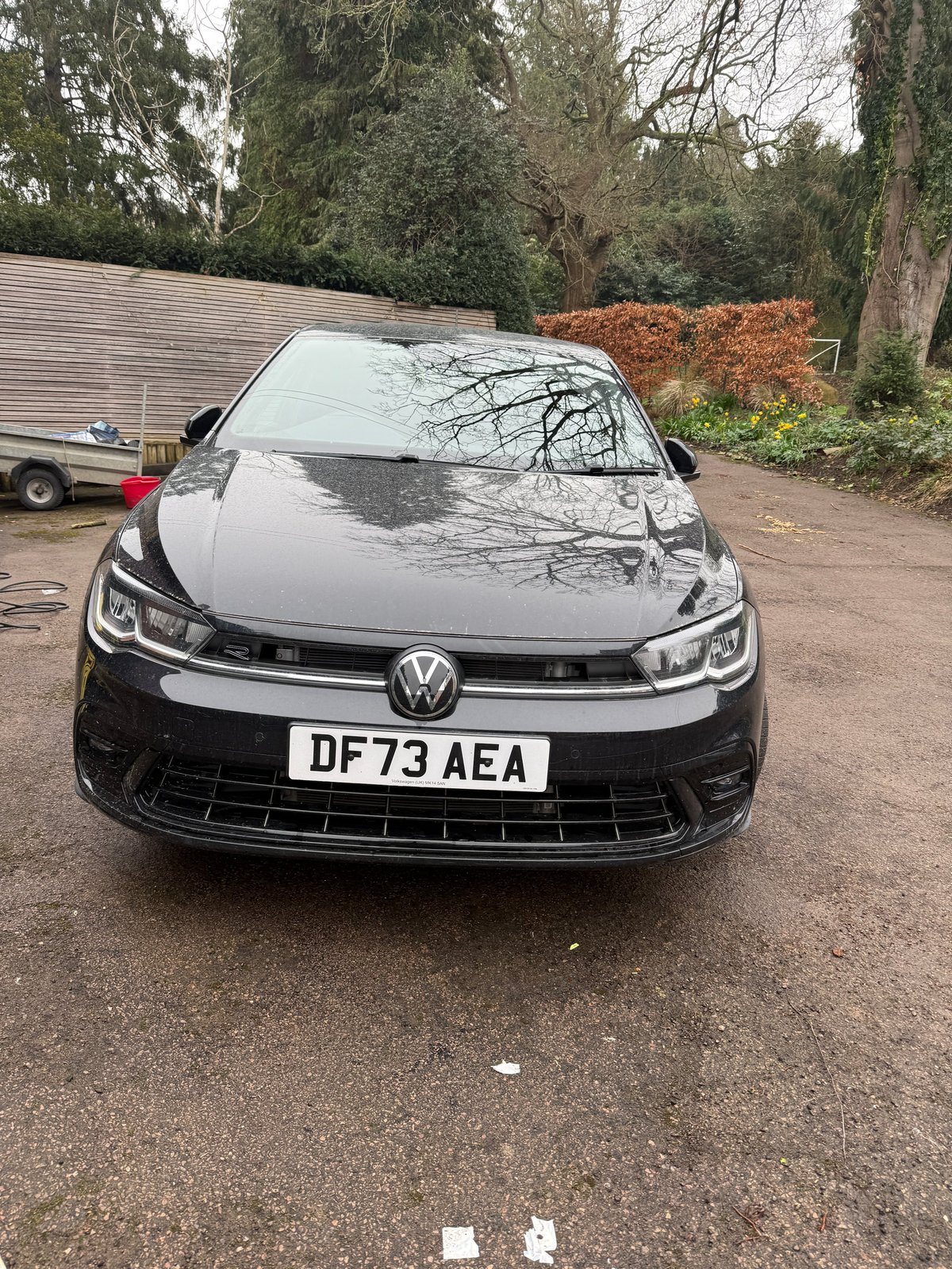 Front view of a black and silver Volkswagen hatchback parked on a driveway with trees and outbuildings in the background