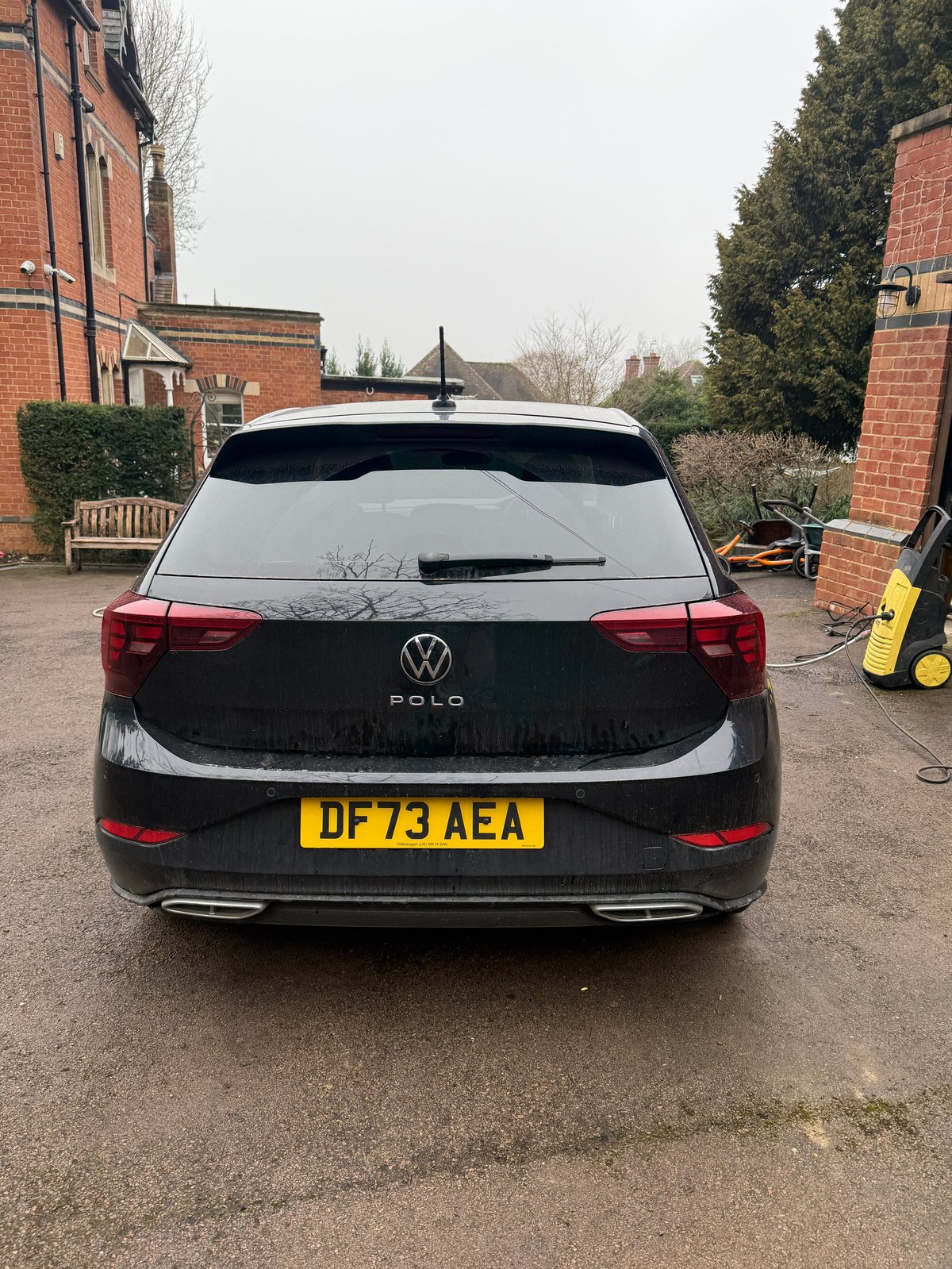 Rear view of a black Volkswagen Polo hatchback parked in a driveway with brick buildings and evergreens visible in the background