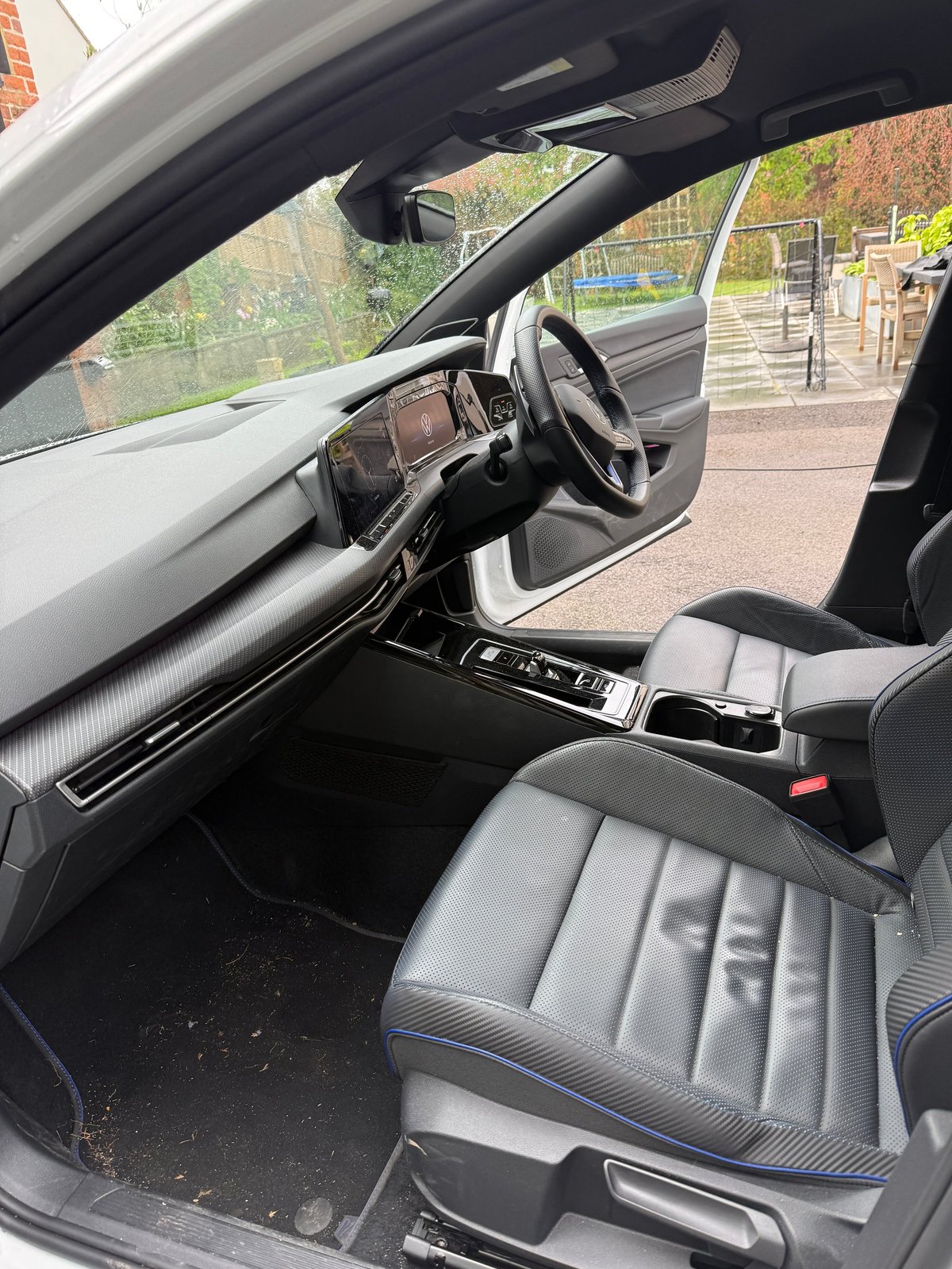 Interior view of a modern car with open drivers door showing steering wheel, dashboard, and gray seats with street and buildings visible outside